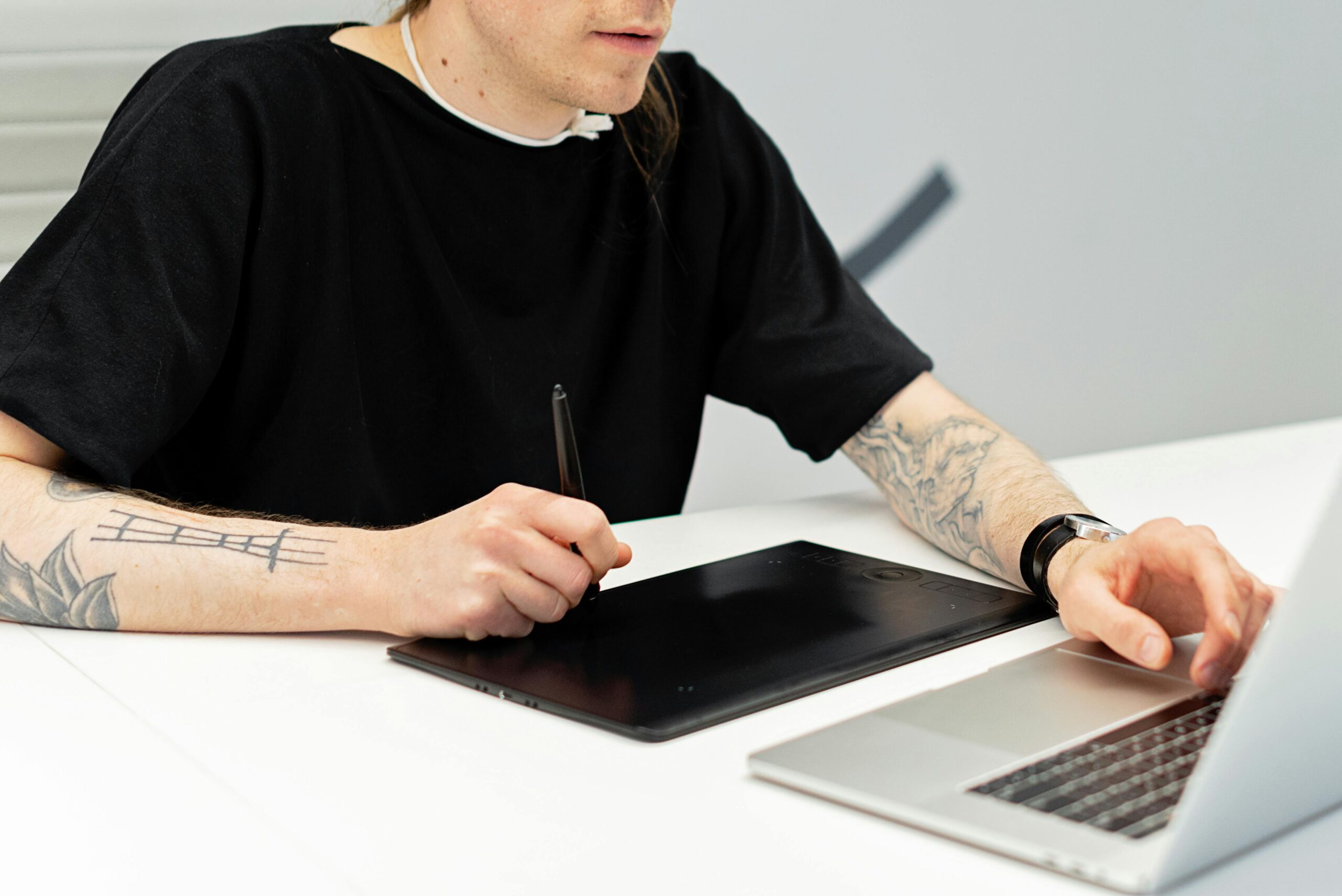 A tattooed artist working on a graphics tablet while using a laptop indoors.